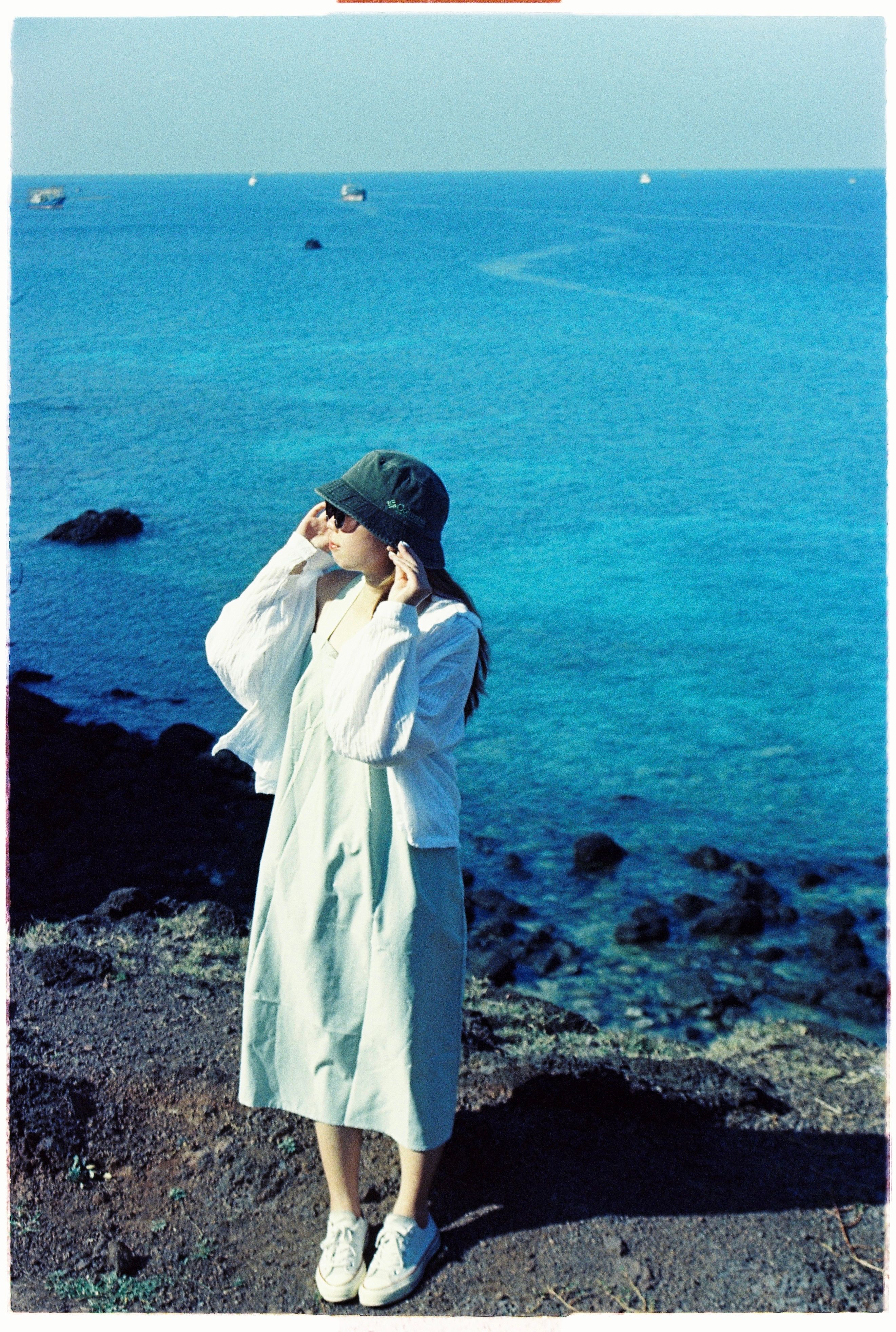 A woman in a hat stands by a clear blue sea on a rocky coastline, basking in sunlight.