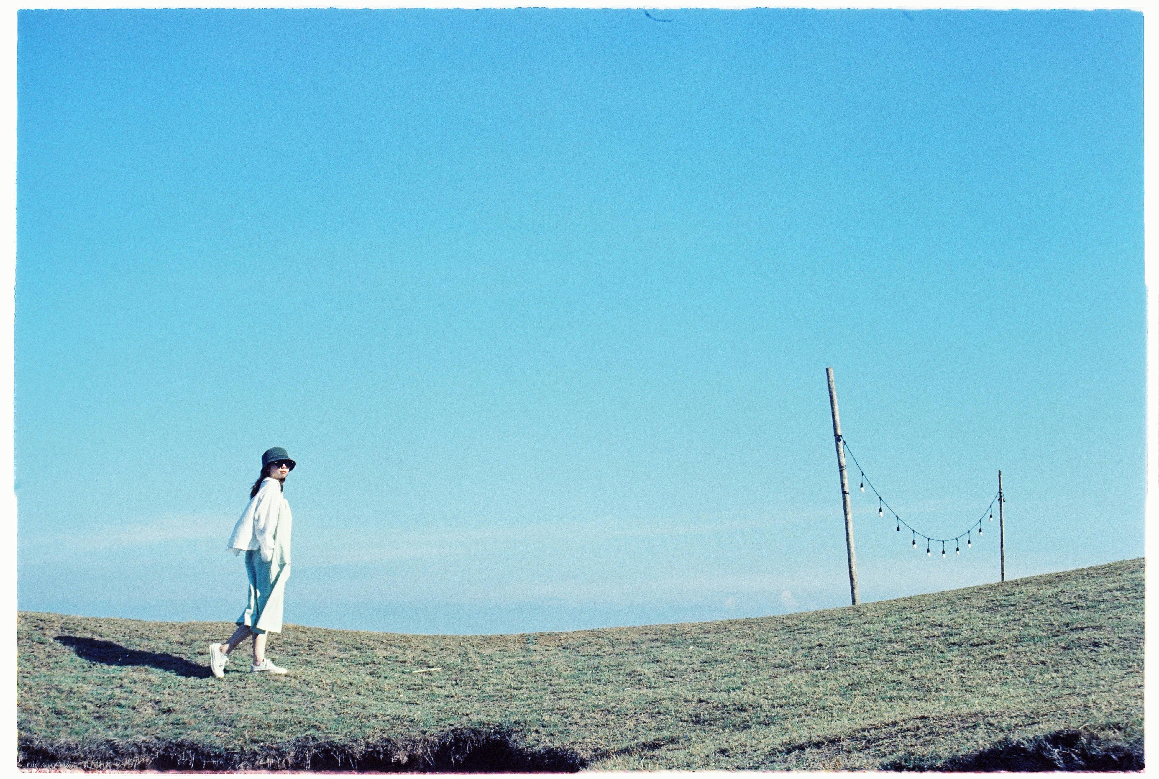 Fashionable woman walking in a serene meadow under a clear blue sky.
