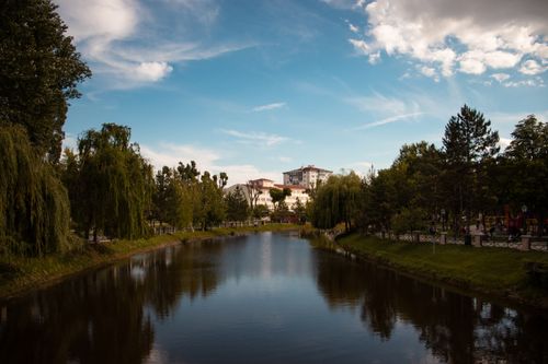 Free A serene park setting featuring a calm lake surrounded by lush trees and distant buildings under a blue sky. Stock Photo