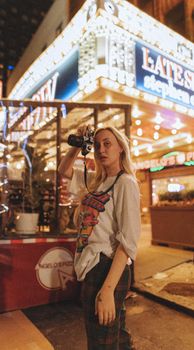 Blonde woman holding camera at night in urban setting with bright lights.