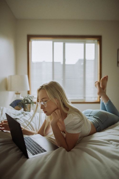 Free A woman in casual attire using a laptop while lying on a bed in a serene and cozy bedroom setting. Stock Photo