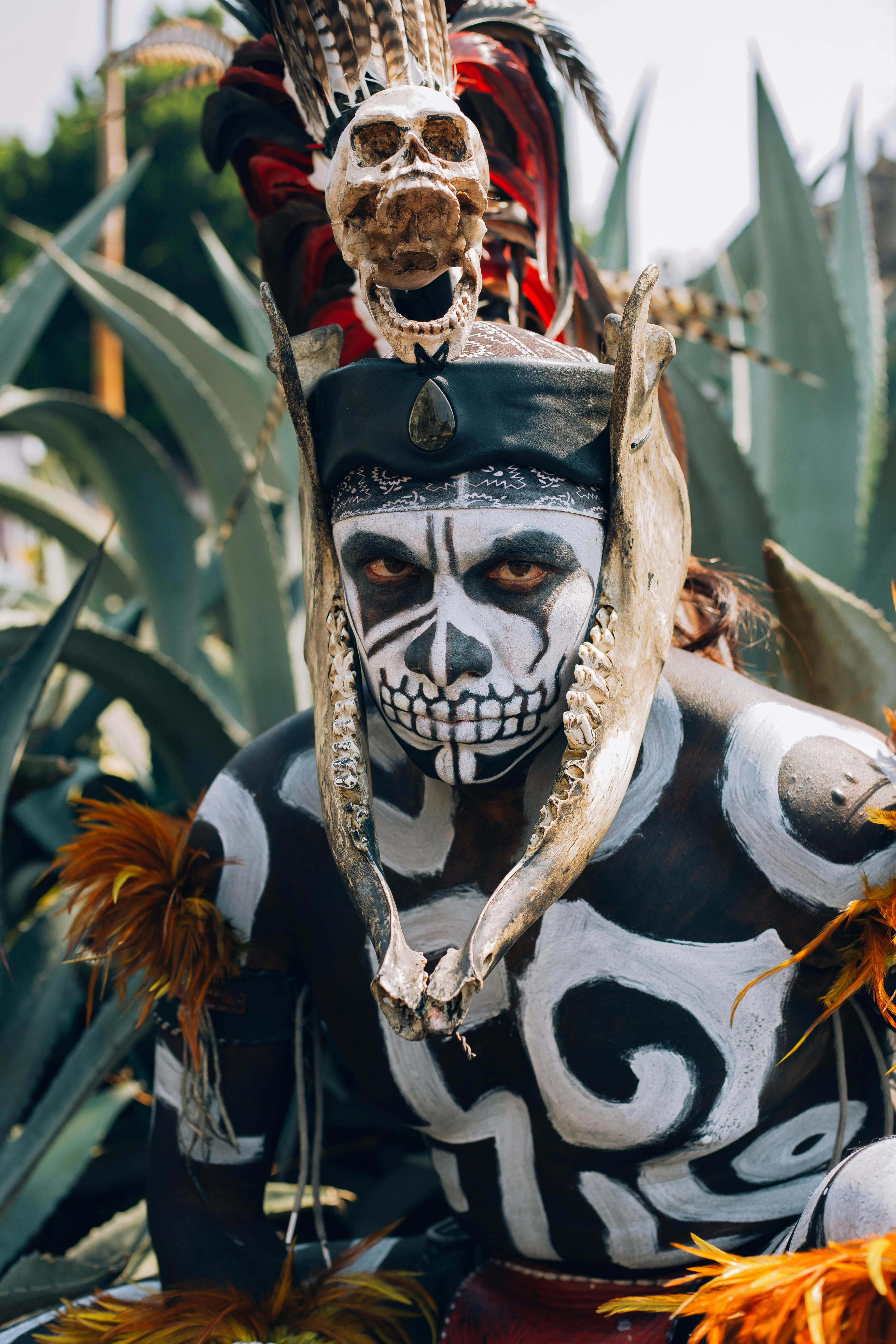 native mexican warrior with pre hispanic makeup simulating a skull and a bone headdress adorned with feathers and cempazuchil flowers