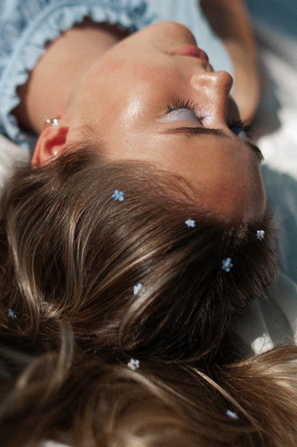 Close-up portrait of a woman lying with small blue flowers in her hair, eyes closed peacefully.