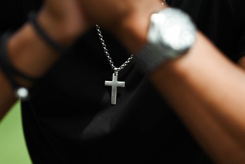 Free A close-up image of a silver cross pendant on a chain necklace worn by a person with a blurred watch in view. Stock Photo