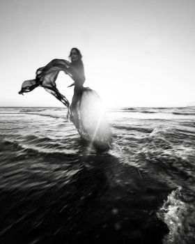Artistic black and white photo of a woman dancing in the ocean at sunset with a flowing scarf.