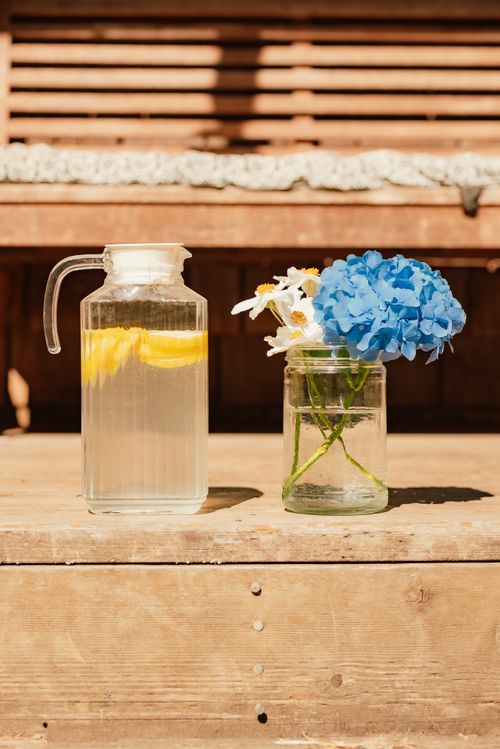 Free Glass pitcher of lemonade with lemon slices next to a jar of fresh blue hydrangeas on a wooden surface. Stock Photo