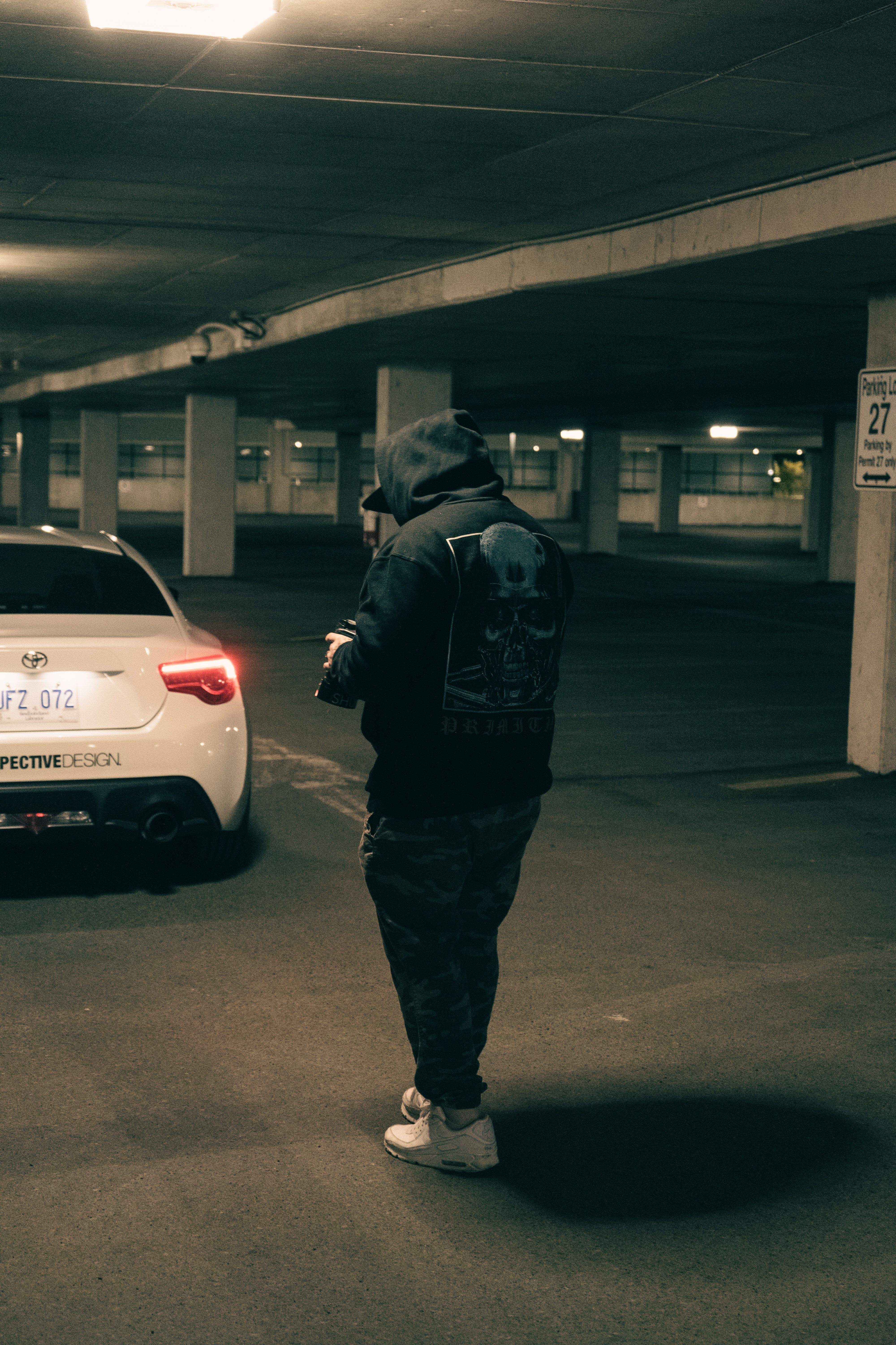 A person in a black hoodie stands near a white sports car in a dimly lit parking garage.