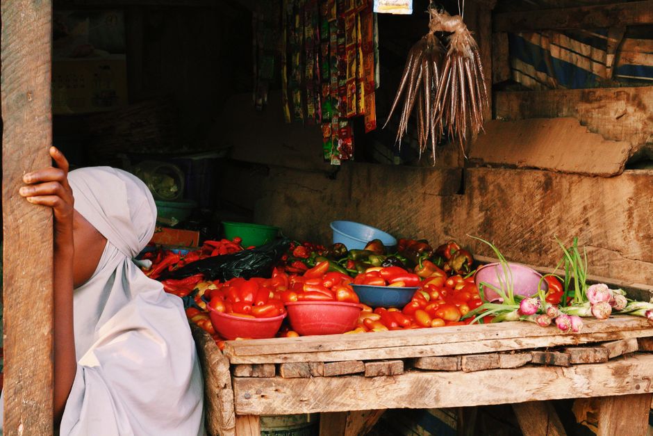 Dark-skinned Nigerian woman in Ankara blouse reviewing her phone screen showing a payment split plan while standing at a busy food market stall in Lagos, natural daylight, photorealistic