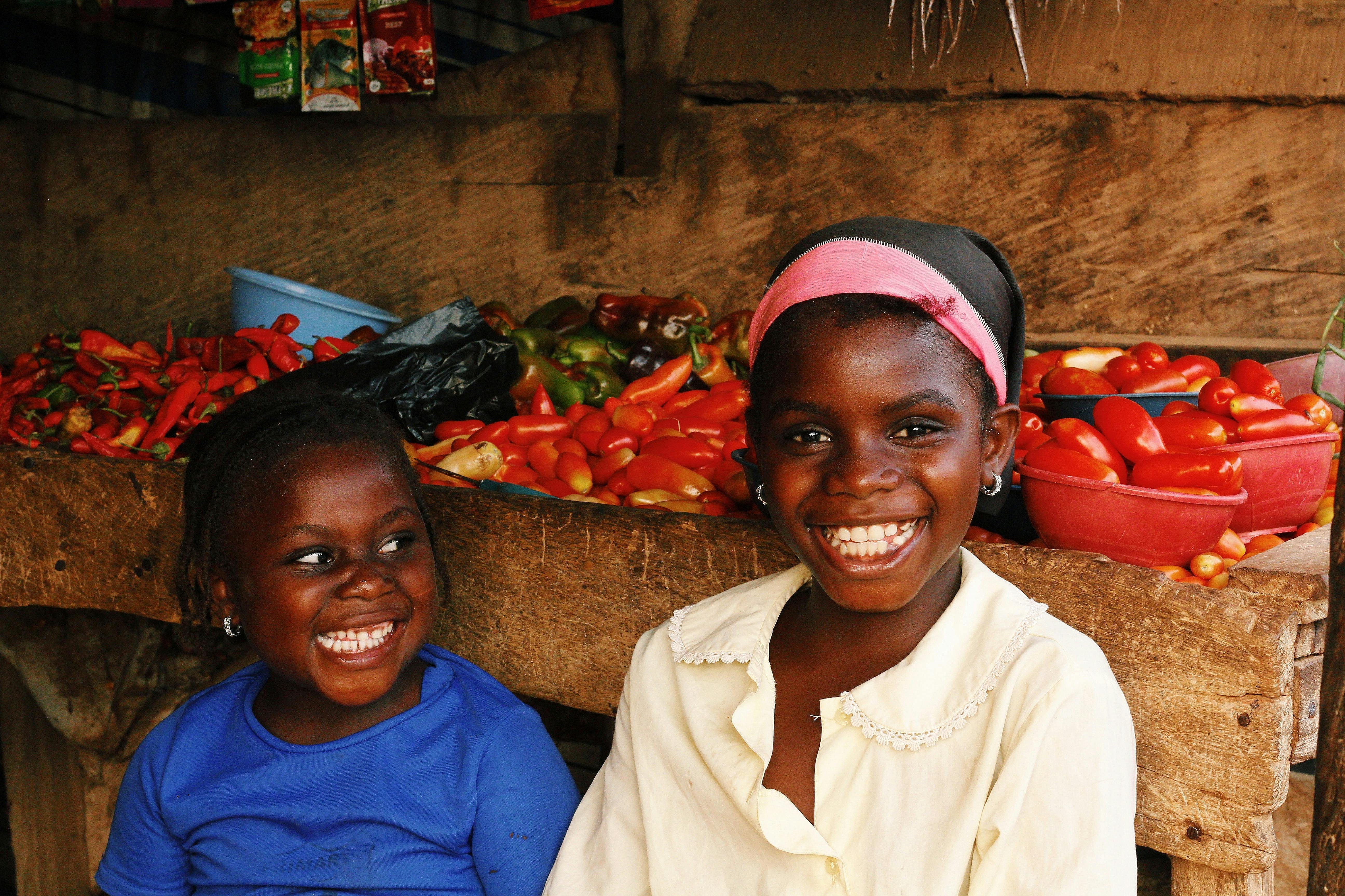 Two African girls smiling in a vibrant market setting with fresh peppers in Abuja, Nigeria.