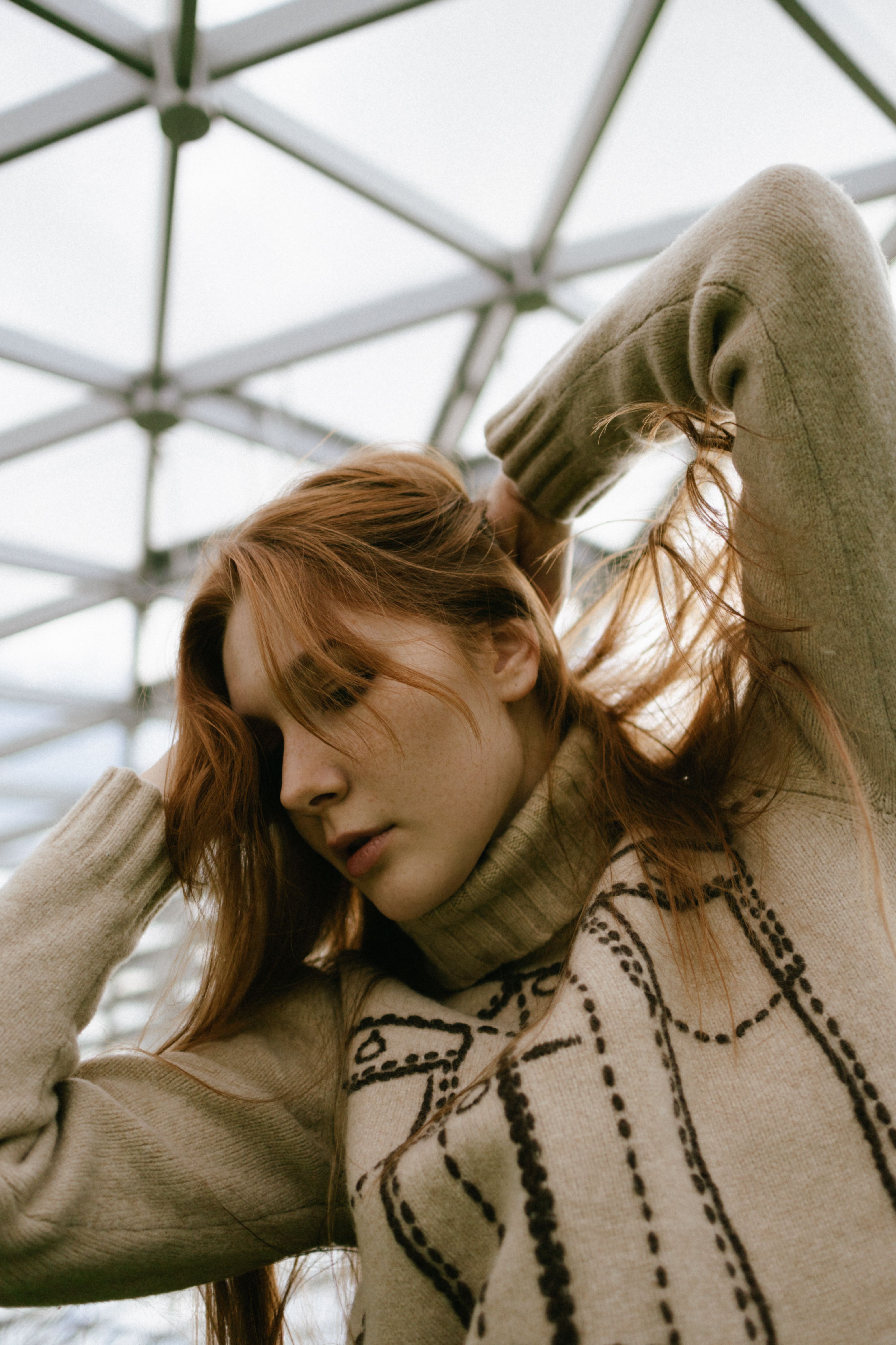 A serene indoor portrait of a redheaded woman adjusting her long hair under a geometric ceiling.