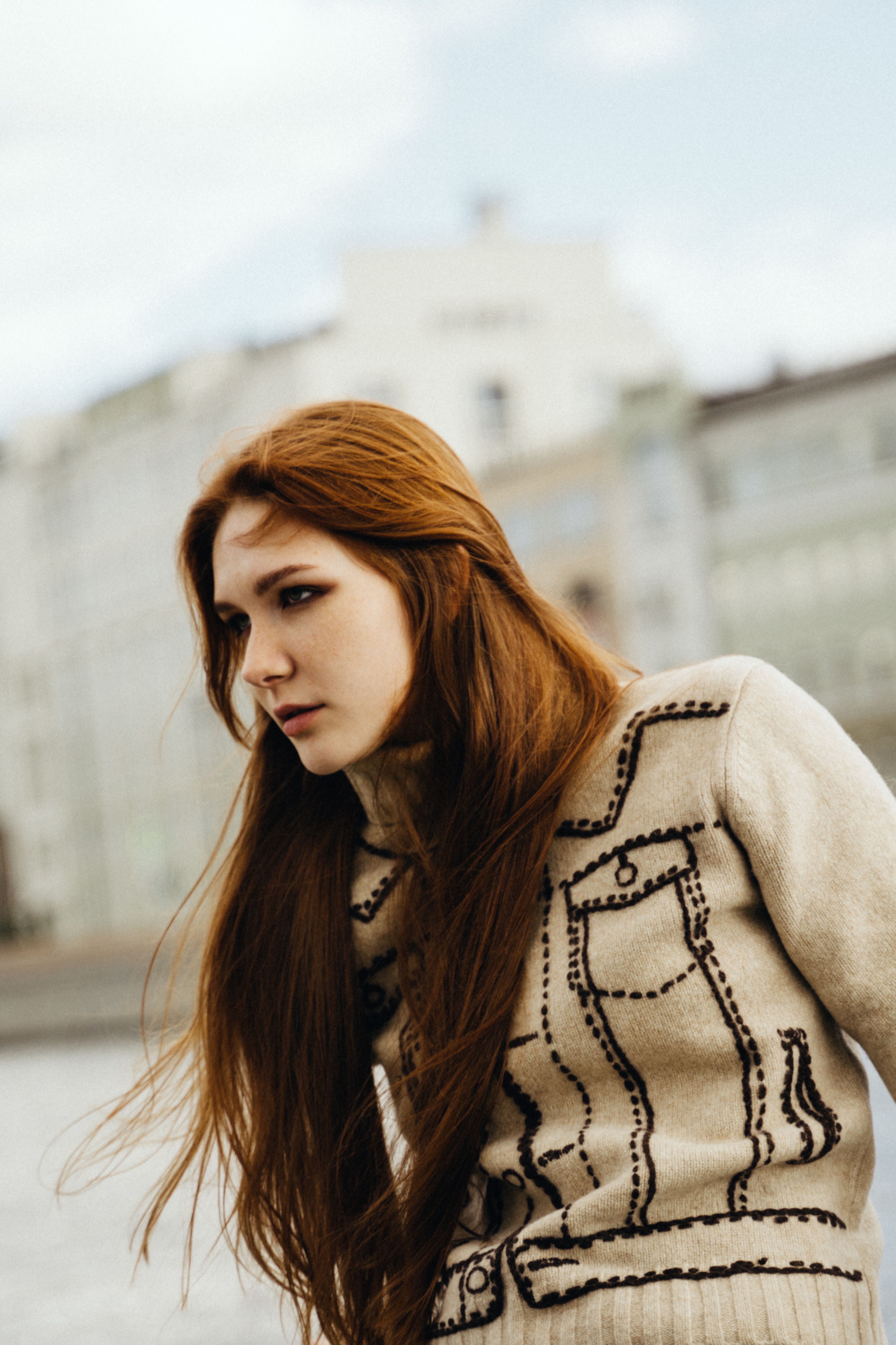 A woman with long red hair in a cozy sweater gazing thoughtfully outside against a city backdrop.