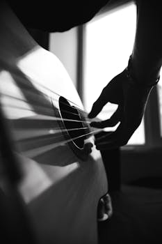 A black and white close-up of a hand playing an acoustic guitar, captured in silhouette against a window light.