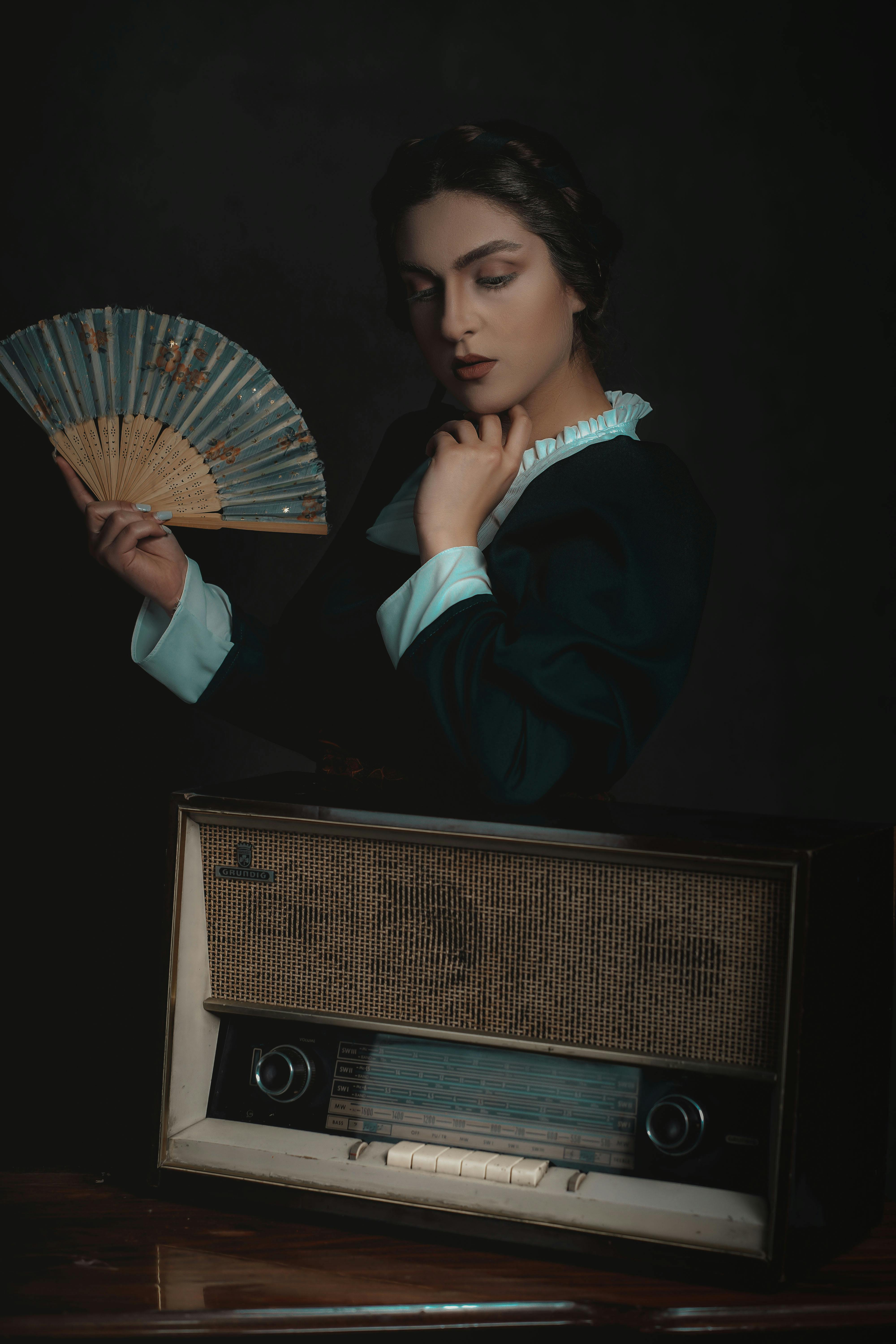 Elegant woman holding a fan poses by a vintage radio. Retro charm, dark tones.