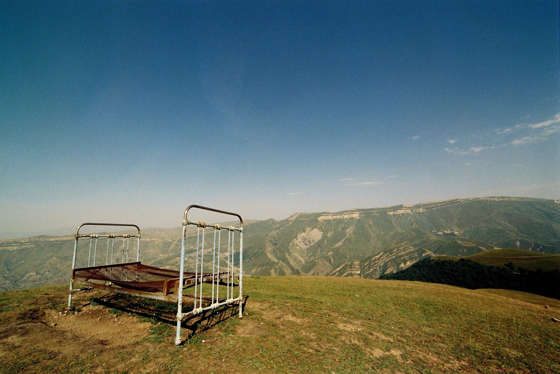 A solitary rusty bed frame stands on a hilltop with a vast mountainous backdrop under a clear sky.