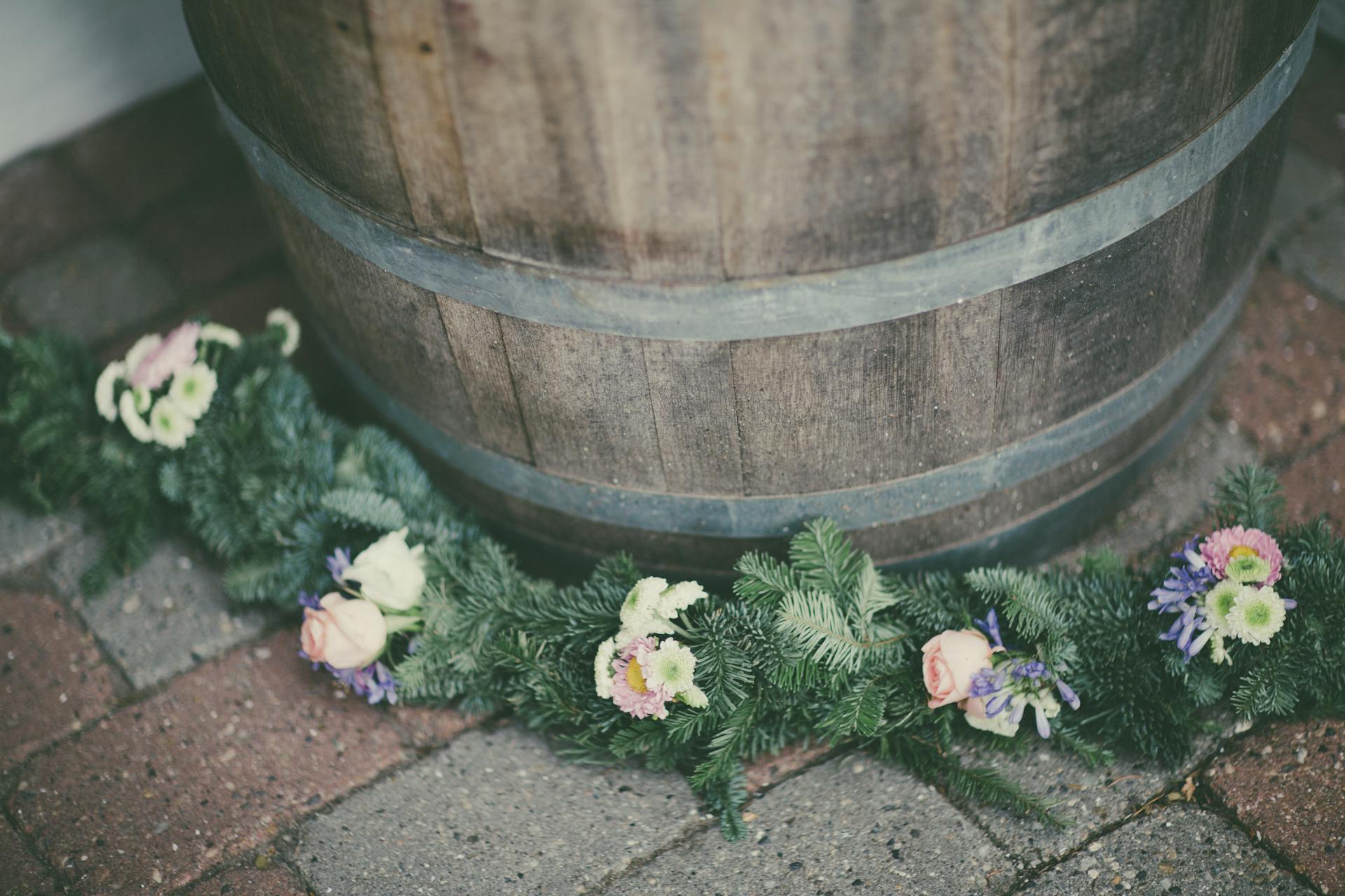 Decorative floral garland draped around a rustic wooden barrel on a brick floor.