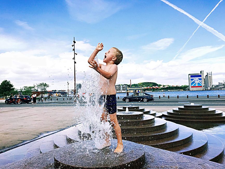 Young boy enjoying a splash in a public fountain on a sunny day by the waterfront.