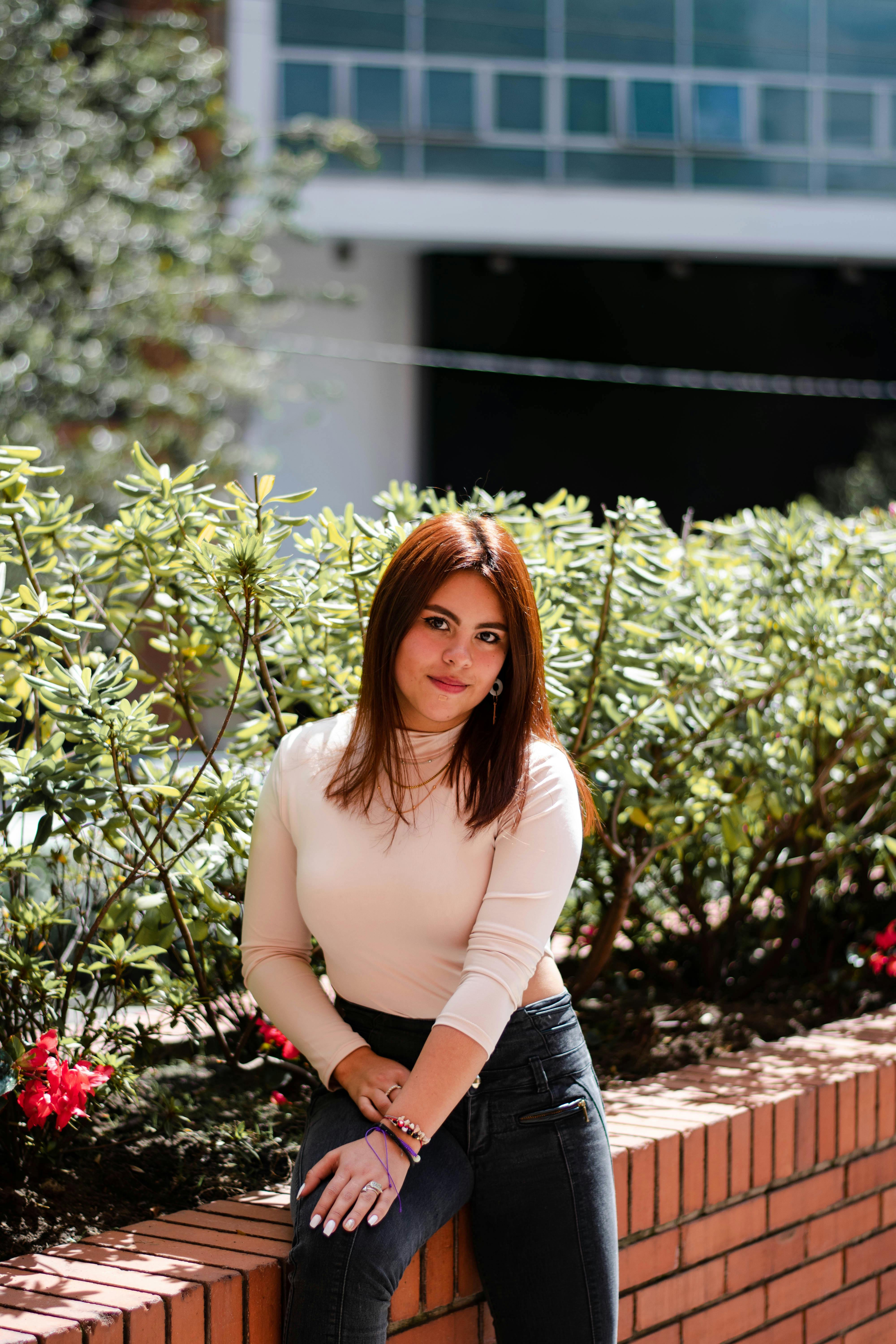 A young woman posing outdoors by a brick wall with greenery, enjoying a sunny day.
