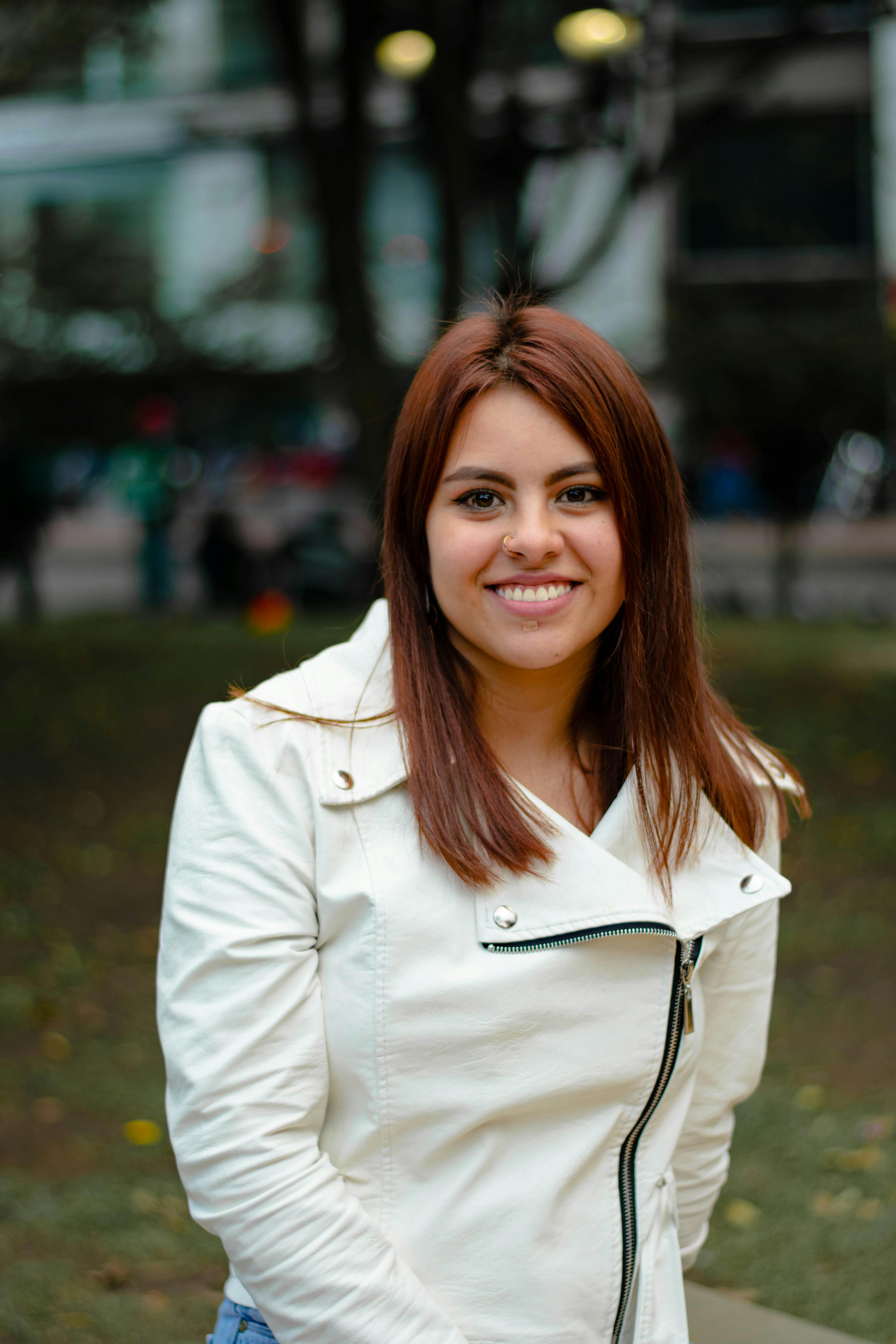 A woman smiling outdoors in a park wearing a white jacket.