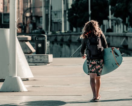 Back view of a surfer carrying a surfboard on a city street, wearing floral shorts and a backpack.