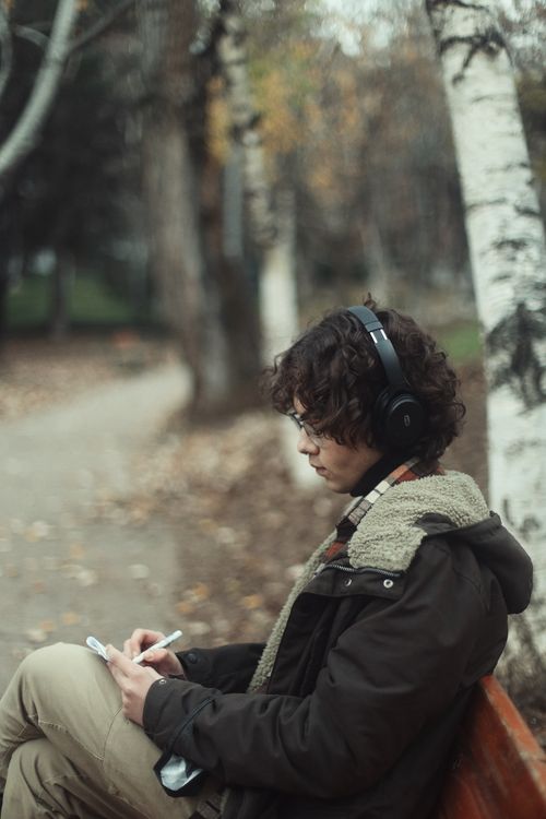 Free Man sitting on a bench in a fall park, wearing headphones and writing. Stock Photo