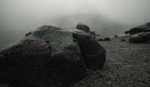 A mysterious coastal scene with large rocks enveloped by thick fog on a misty shore.