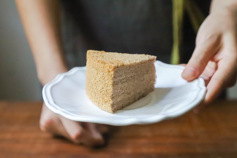 Hand holding a piece of homemade cake on a white plate, offering it as a treat.