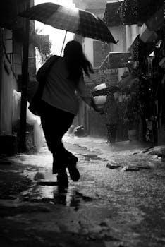 A black and white image of a woman walking down an alley with an umbrella on a rainy day.