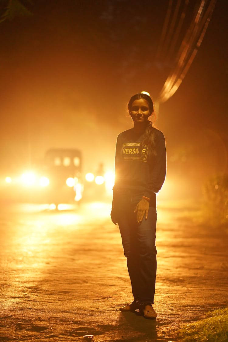 Girl Standing On The Road At Night With Car Lights Behind Her 