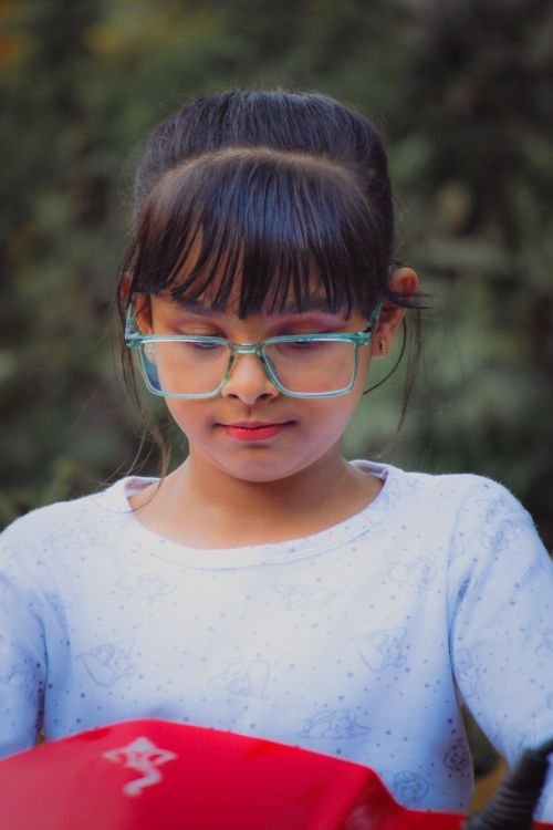 Free Portrait of a young girl with glasses, outdoors, focusing on a red object. Captured in Madikeri, India. Stock Photo