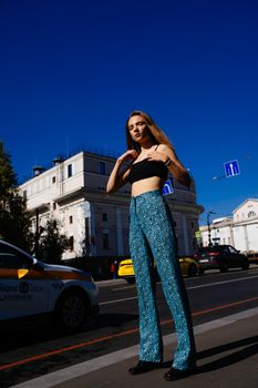 Confident woman wearing blue patterned pants poses on a vibrant city street in bright sunlight.