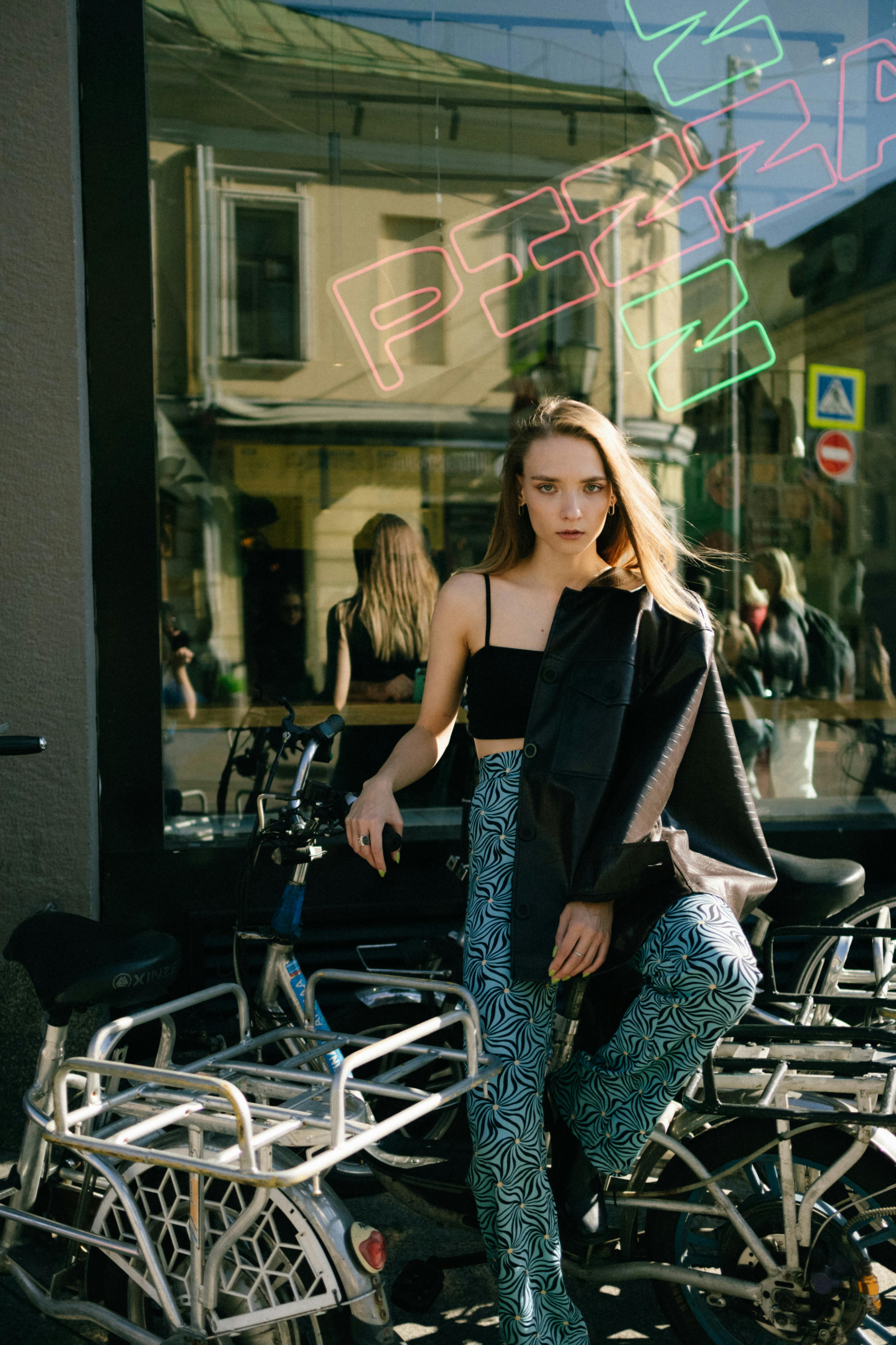 Trendy woman in stylish outfit posing with bicycle outside pizza restaurant.