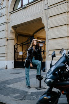 Stylish woman in leather jacket posing by motorcycle in urban setting.