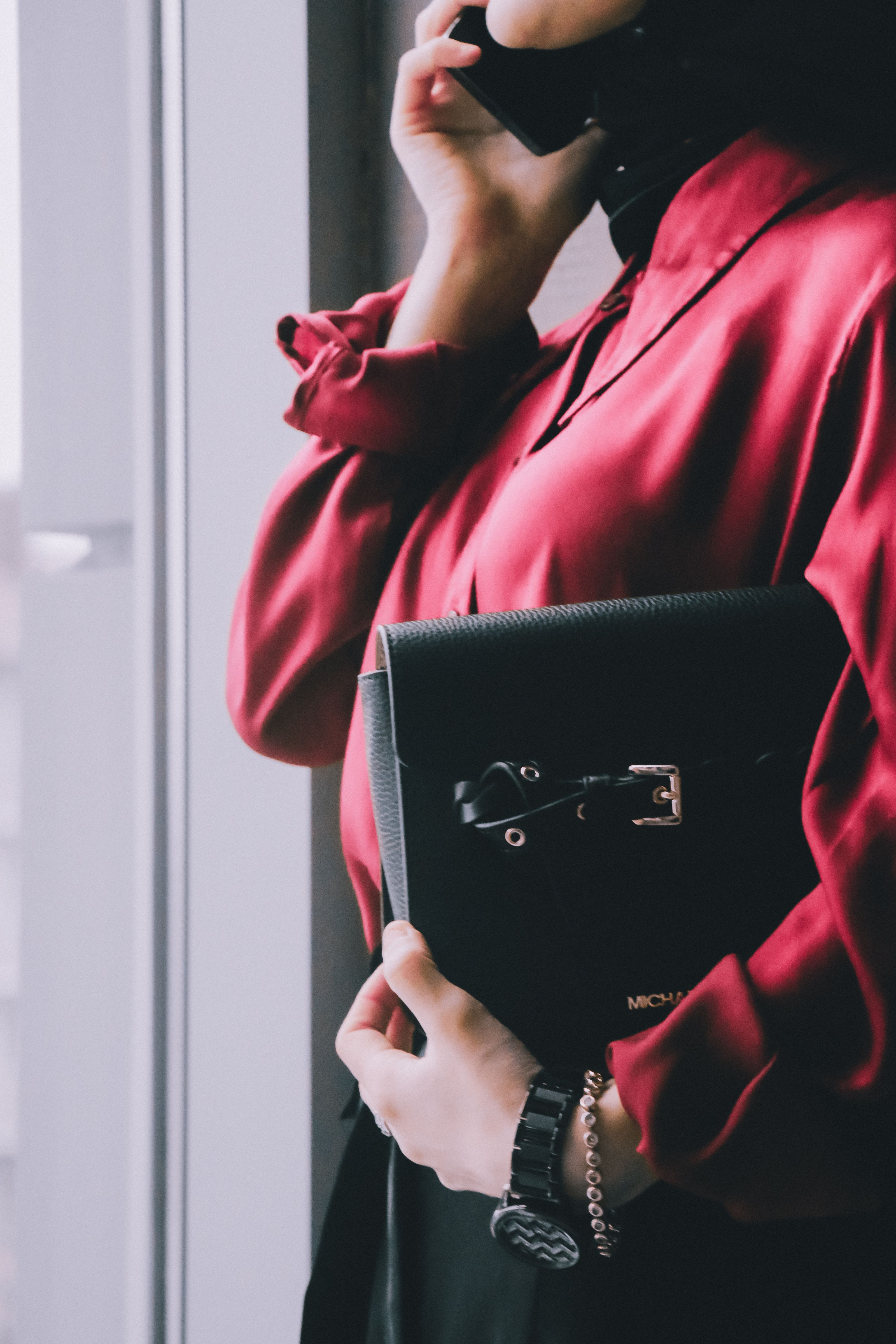 Mulher sofisticada com camisa vermelha segurando clutch de couro preto ao lado da janela, exalando elegância.