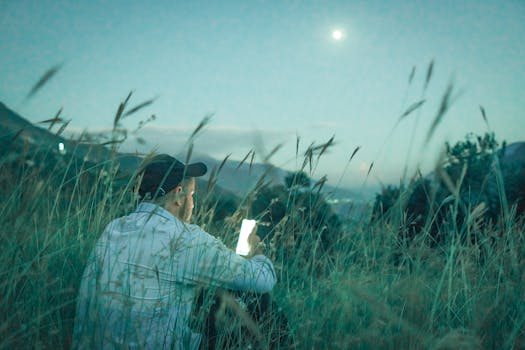 A man sits in a grassy field at night, illuminated by his smartphone under a moonlit sky.