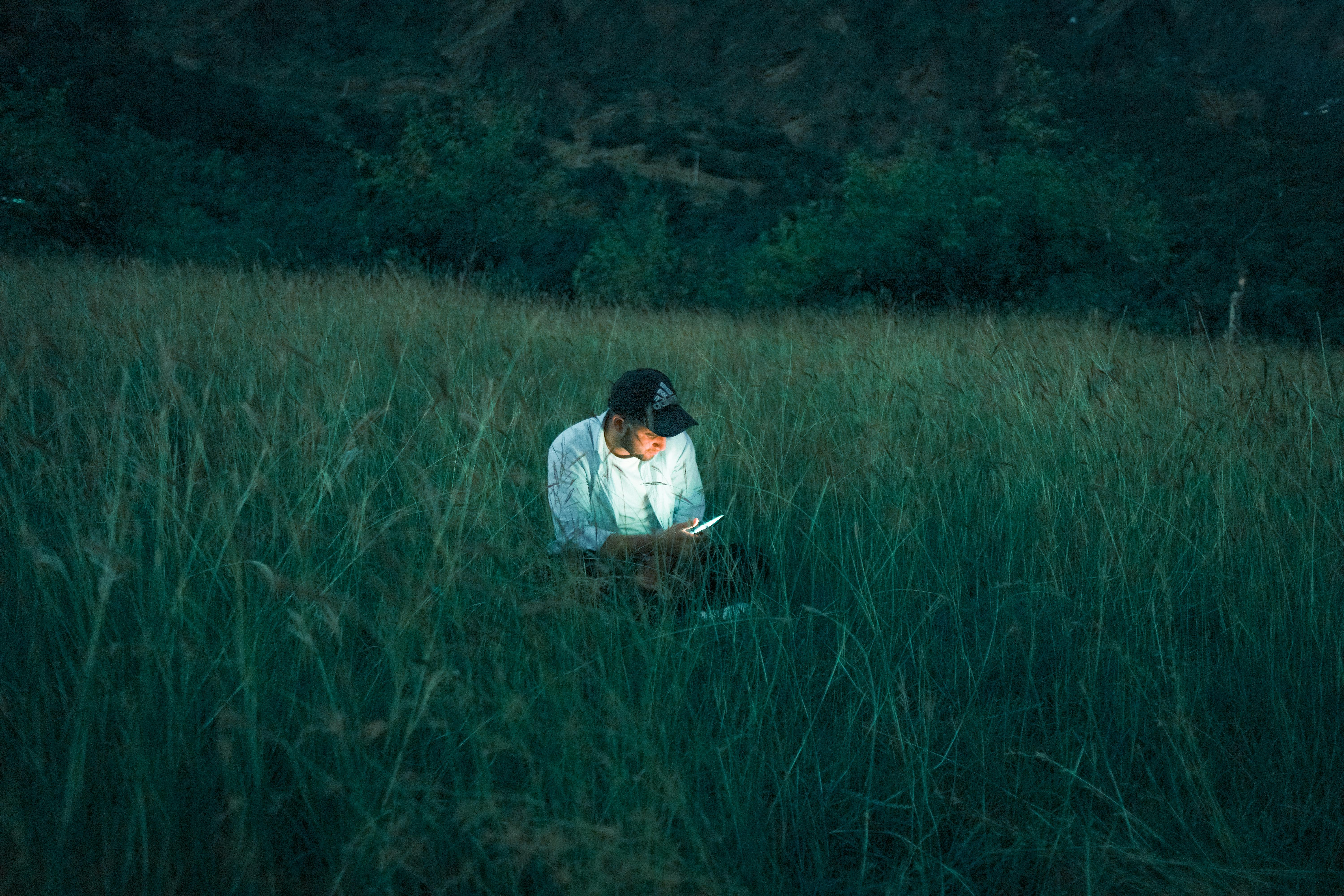 A man sitting in a grassy field using a smartphone during twilight.