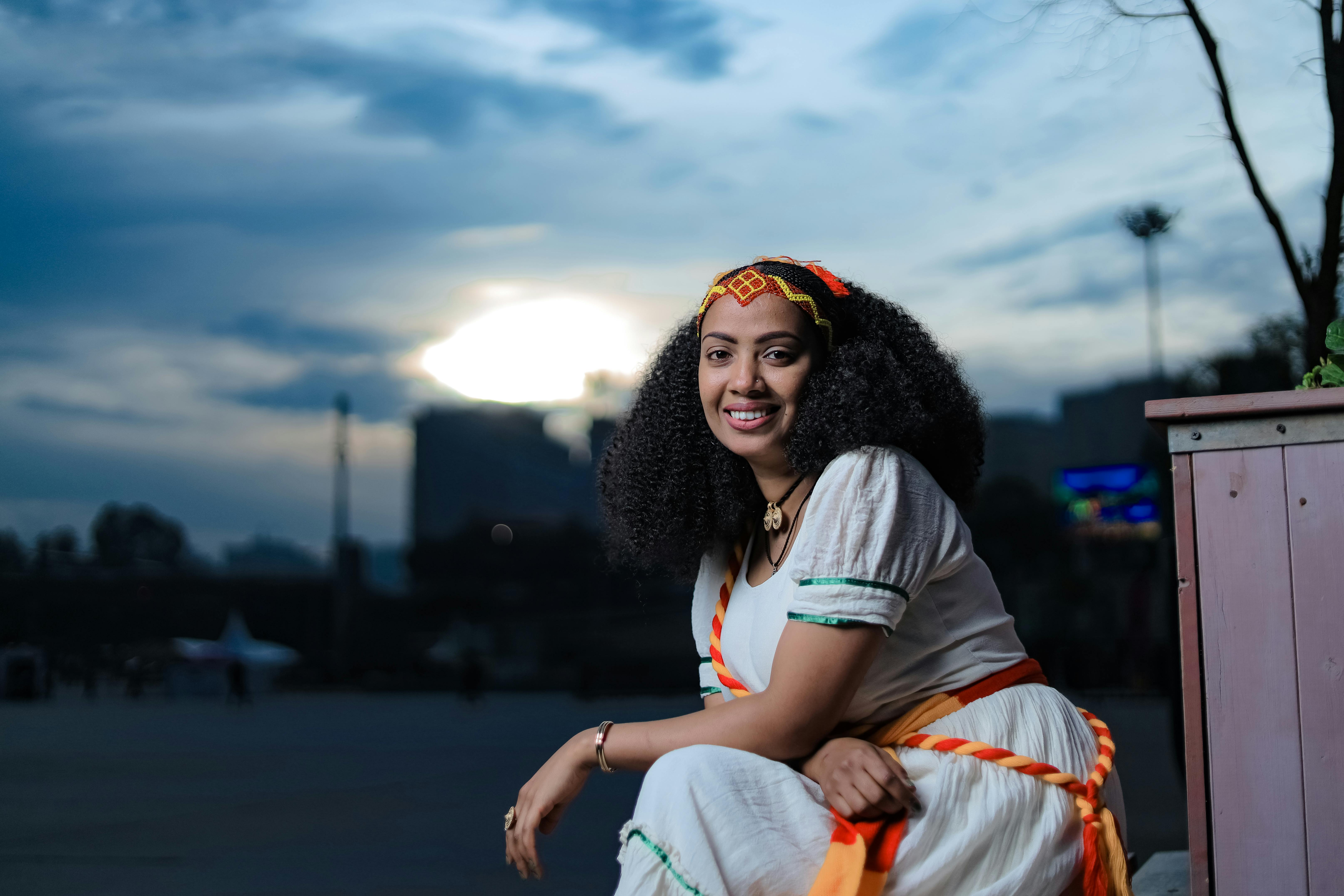 Portrait of a smiling Ethiopian woman in traditional attire, seated outdoors in Addis Ababa at twilight.