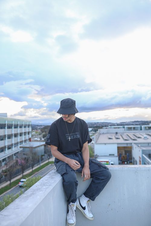 Free Stylish young man in casual wear sitting on a rooftop edge under a cloudy sky. Stock Photo