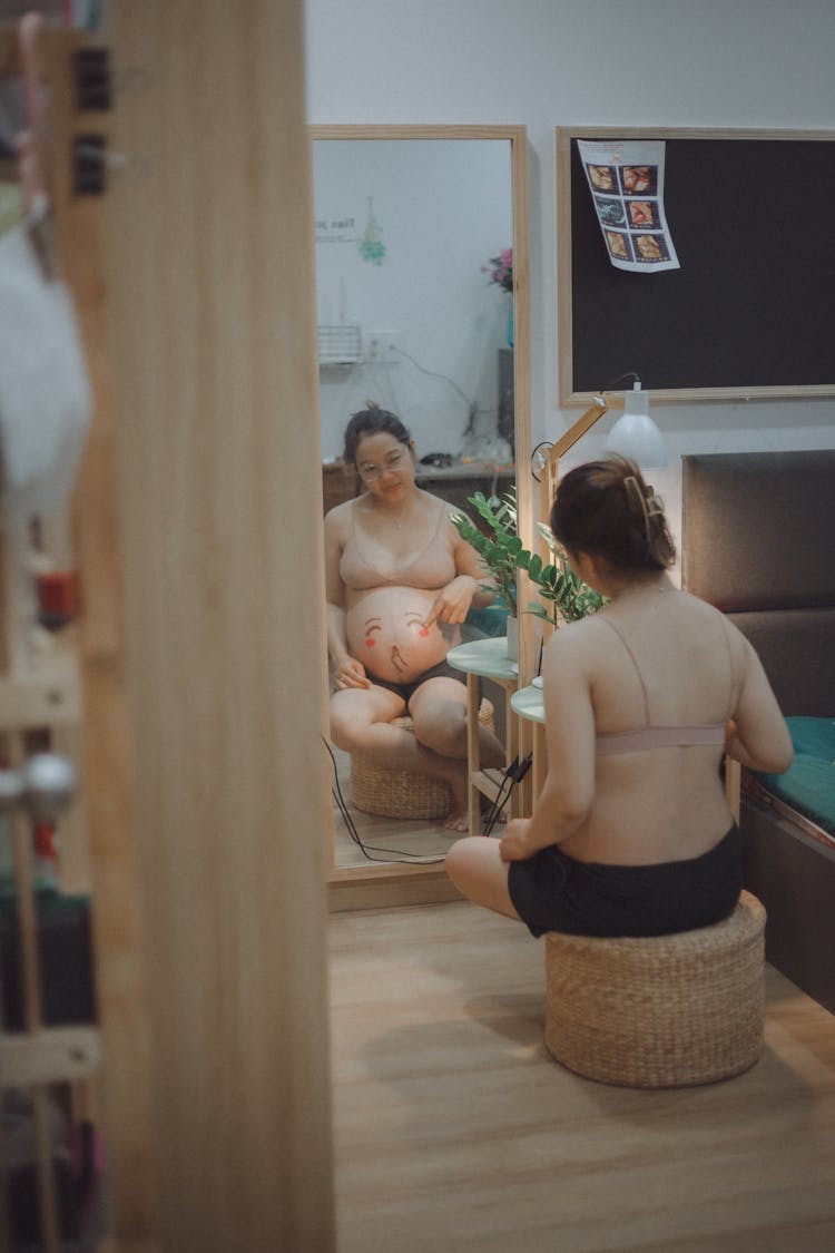 A Pregnant Woman Sitting On A Woven Chair While Looking At Herself In The Mirror