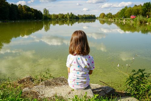Free A child sits by a peaceful lake, enjoying the serene natural surroundings. Ideal for outdoors and tranquility themes. Stock Photo