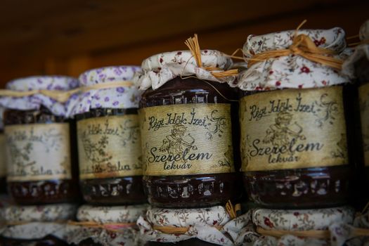 Close-up of rustic homemade jam jars with floral fabric covers and rustic labels on a wooden shelf.