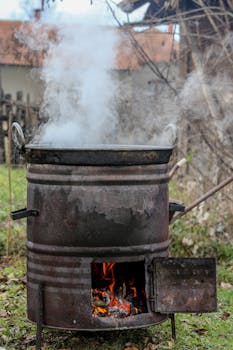 A steaming pot over a rustic wood stove outdoors emitting smoke and flames.