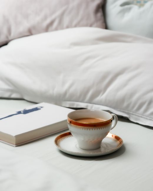 A warm cup of coffee on a saucer beside a book on a cozy bed with soft pillows.