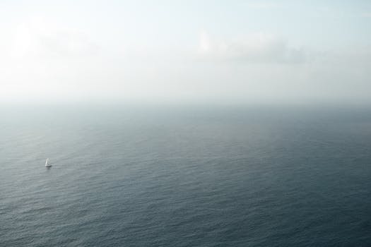 A peaceful aerial capture of a lone sailboat on a calm, foggy ocean under a cloudy sky.