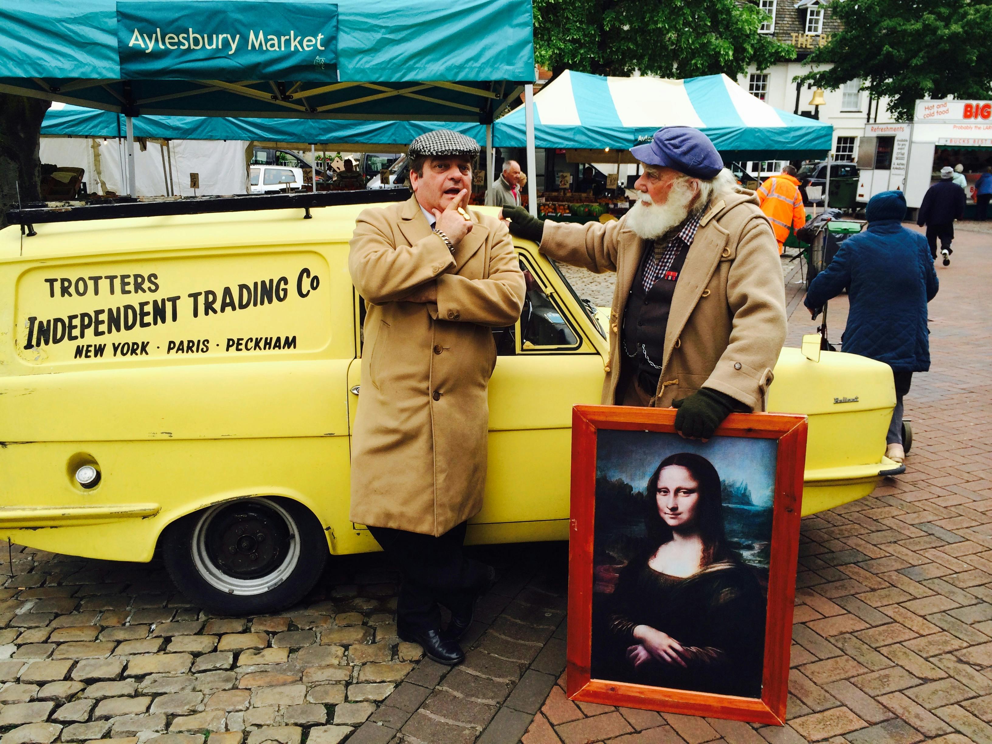 Two men in vintage attire at Aylesbury Market with a classic yellow car and Mona Lisa painting.