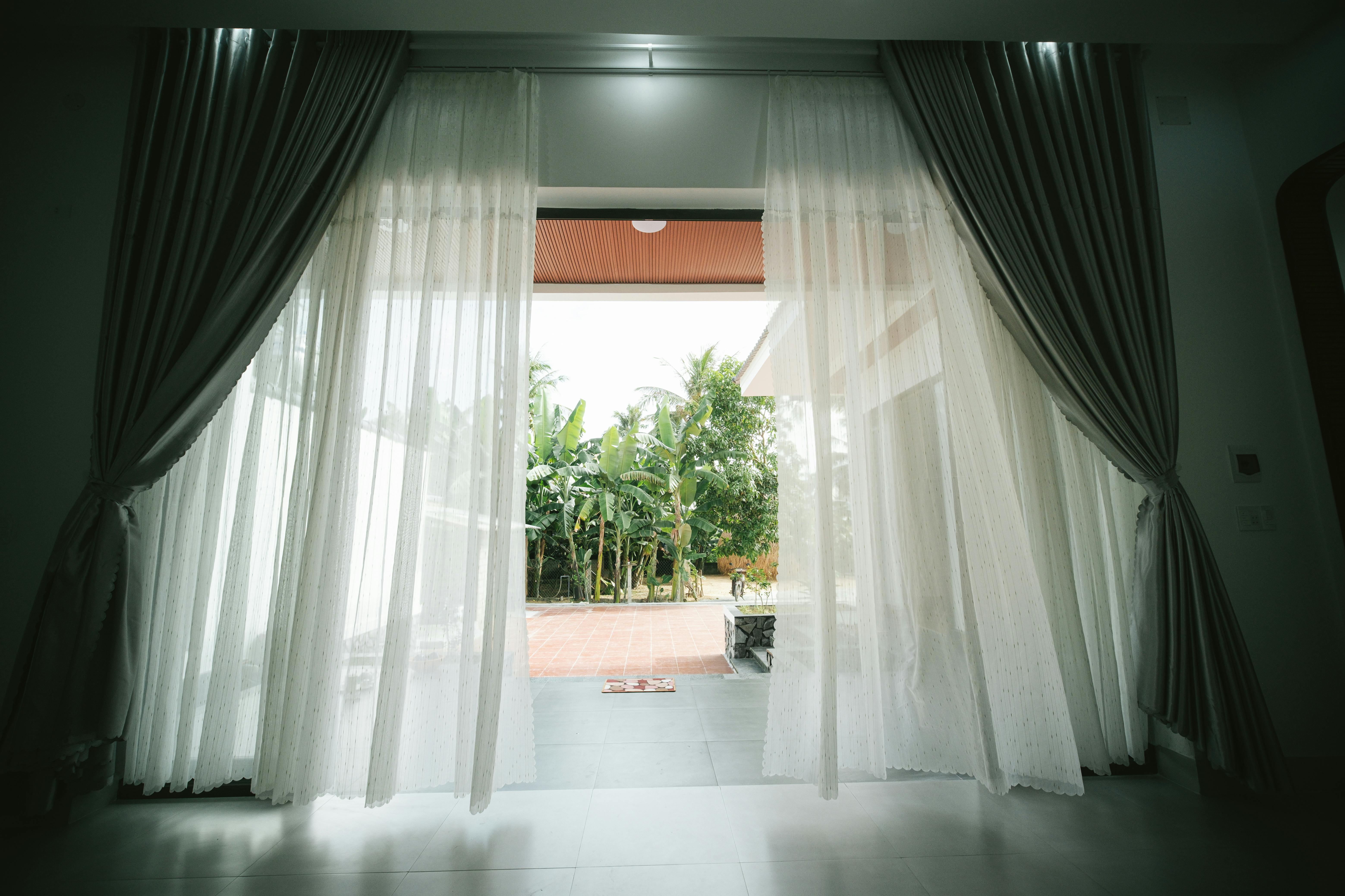 View of open terrace through white curtains, revealing a lush garden.