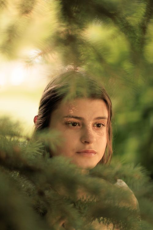 Free Beautiful close-up portrait of a young woman peeking through lush green foliage in a serene natural setting. Stock Photo