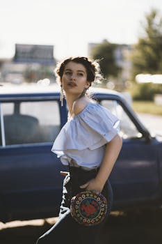 阳光明媚，一位身穿时尚露肩装的年轻女子倚在一辆老式蓝色汽车旁摆姿势，尽显都市别致风范