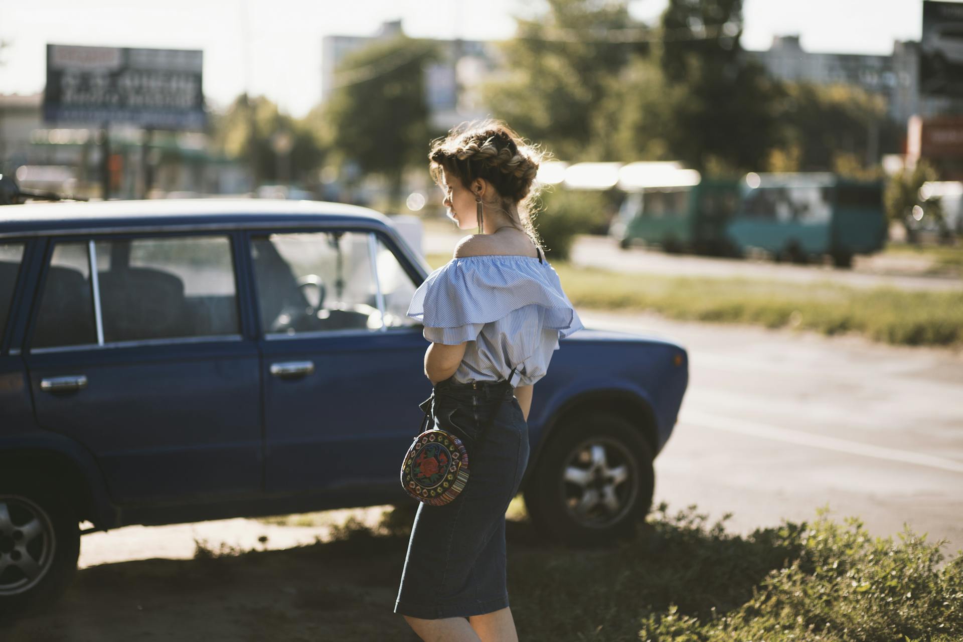 A fashionable woman stands by an old blue car in a sunny outdoor setting.