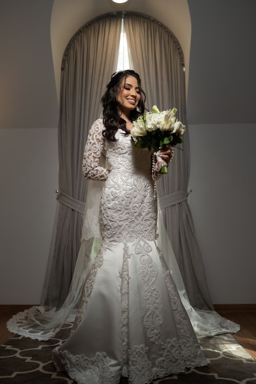 Free Stunning bride smiles holding bouquet in a decorative lace wedding gown indoors. Stock Photo