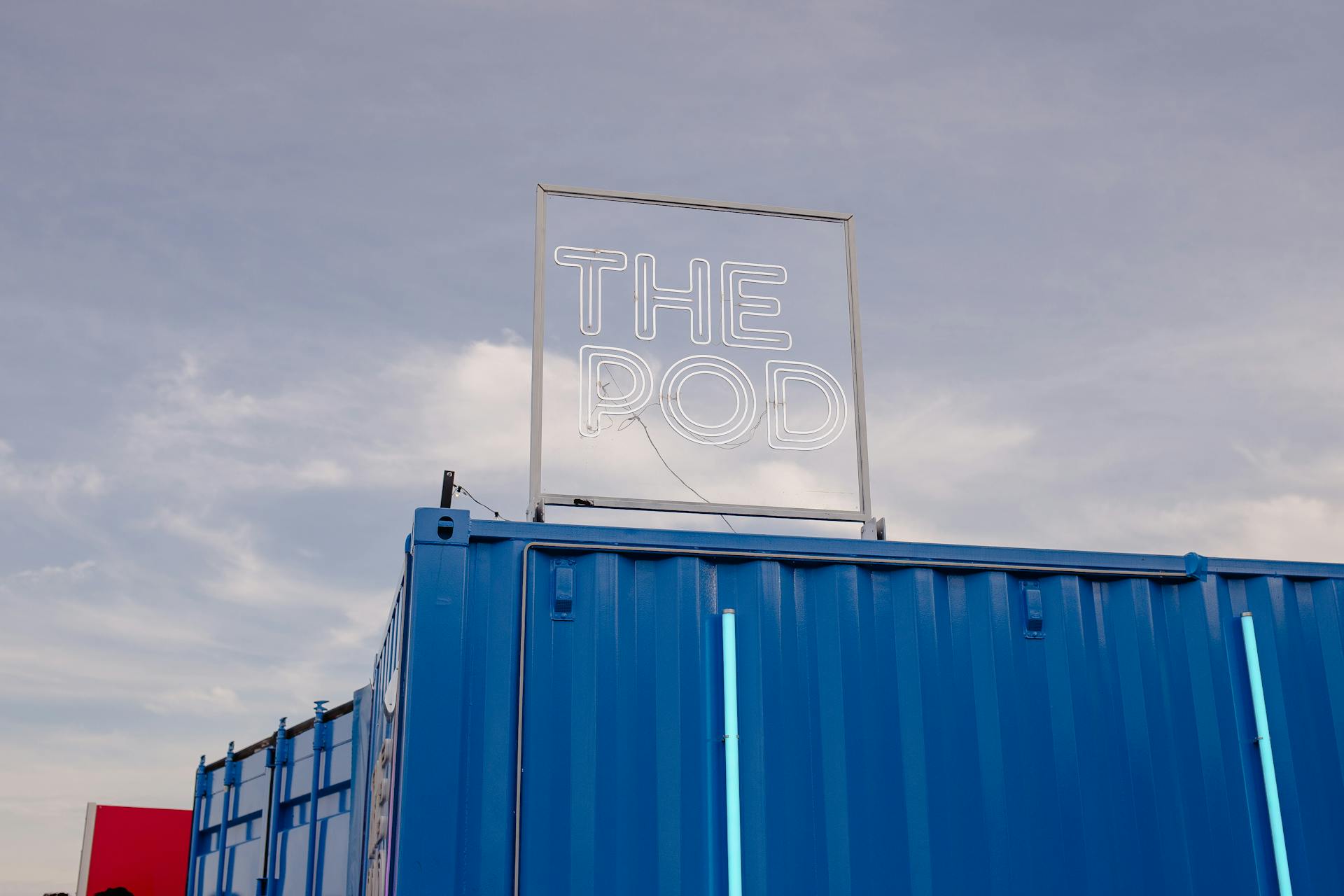 Blue shipping container with 'The Pod' neon sign under a clouded sky.